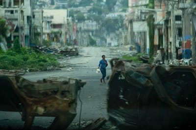 na persona pasa junto a vehículos incendiados por bandas armadas —utilizados como barricada durante los enfrentamientos de la semana pasada con las fuerzas de seguridad haitianas— en una calle desierta del centro de la ciudad; imagen captada desde un vehículo policial blindado durante una patrulla en Puerto Príncipe, el 16 de enero de 2026. AFP